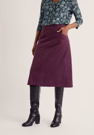 Woman wearing a purple knee-length skirt with pockets, black knee-high boots, and a blue floral long-sleeve blouse standing against a neutral background.