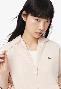 Sudadera con cremallera de color rosa claro, fabricada con tejido texturizado y detalle de cordón. Presenta un pequeño logo de cocodrilo verde en el pecho.