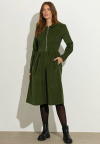 Groene corduroy jurk met lange mouwen, kraag en een ritssluiting aan de voorkant. Heeft plooien en zijzakken. Gekozen met zwarte enkelboots.