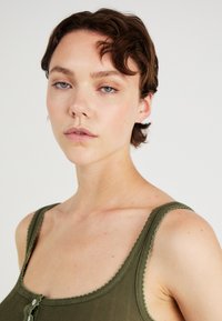 Green ribbed tank top with scalloped edges, featuring a button placket at the front and a fitted silhouette. Model has short hair.