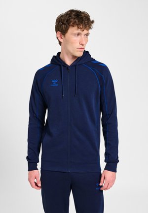 Mężczyzna w granatowej kurtce z zamkiem i kapturem oraz pasujących spodniach z niebieskim logo i akcentami, patrzy w lewo.