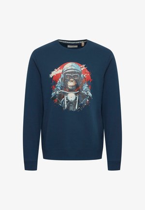 Tume sinine spordipusa, millel on graafika kiivris ahvist, kes sõidab mootorrattaga, ja pritsitud punased ja valged aktsendid.