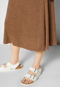 Sandálias brancas Birkenstock com duas tiras ajustáveis e fivelas de metal, usadas com uma saia castanha canelada num fundo neutro.