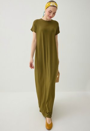 Vrouw draagt een losse maxi jurk in olijfgroen, gele hoofddoek, mosterdkleurige platte schoenen en houdt een kleine gevlochten handtas vast, staande tegen een effen achtergrond.