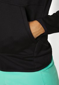 Main partiellement dans la poche avant d'un sweat à capuche noir porté avec un pantalon vert clair sur fond blanc.