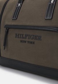 Sac de sport en toile vert foncé avec des accents en cuir noir, présentant deux poignées robustes, une poche zippée à l'avant et un logo en relief.