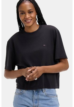 Femme souriante aux longues tresses, portant un T-shirt noir court et un jean bleu clair, les mains jointes devant, sur un fond blanc.