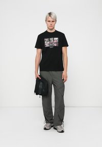 Sort t-shirt med grafisk print, grå cargo-bukser og grå sneakers. Modellen holder en lille sort taske. Ren baggrund, neutral belysning.