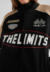 Černá a béžová bunda s závodními nášivkami, včetně nápisů "RACE ON", "LIVE LIFE LIKE IT'S THE LAST TAP", zkřížených šachovnicových vlajek a "THE LIMITS".