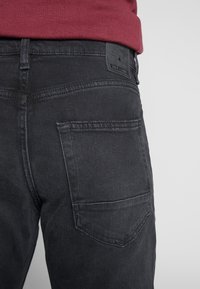 Musta farkkujen takatasku näkyvissä, teksturoitu pinta. Vyötäröllä musta nahkainen brändilaatta.