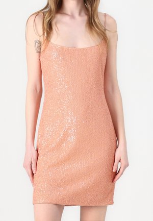 Femme portant une mini-robe sans manches, pailletée de couleur pêche avec de fines bretelles, debout sur un fond clair uni.