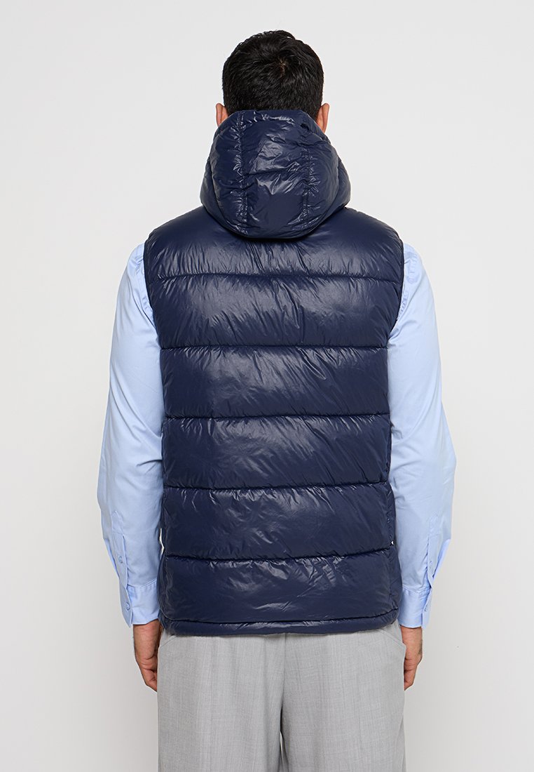 Gilet matelassé bleu marine avec capuche, présentant des sections quiltées et une texture brillante. Porté par-dessus une chemise bleu clair.