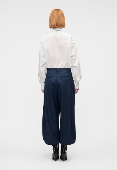 Λευκό πουκάμισο με κουμπιά και μακριά μανίκια, συνδυασμένο με σκούρο μπλε ριγέ παντελόνι φαρδιάς γραμμής και μαύρα μυτερά μποτάκια αστραγάλου.