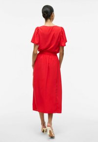 Robe midi rouge avec de courtes manches bouffantes, taille froncée et texture lisse. Portée avec des sandales à talons blocs de couleur claire.