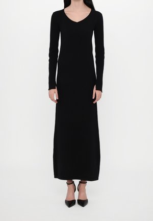 Woman wearing a long black V-neck dress with long sleeves and black pointed-toe ankle strap heels standing against a white background.