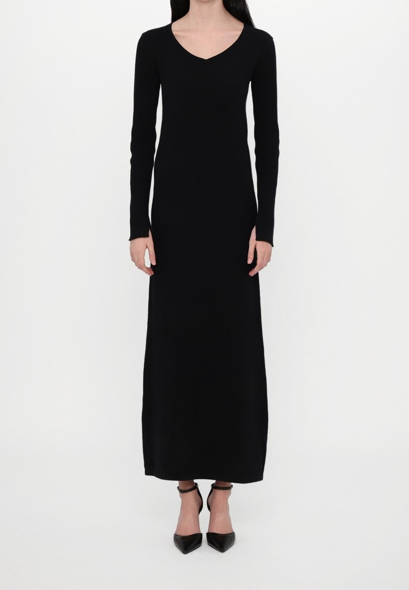 Femme portant une longue robe noire à col en V avec des manches longues et des talons noirs à bout pointu avec bride à la cheville, debout sur fond blanc.