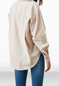 Donna che indossa una camicia beige a maniche lunghe eampie con polsini abbottonati e jeans skinny blu, vista di schiena.