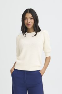 Frau mit langen dunklen Haaren, die einen cremefarbenen, strukturierten Kurzarmpullover und blaue, hoch taillierte Hosen trägt, Hände in den Taschen, leicht lächelnd.