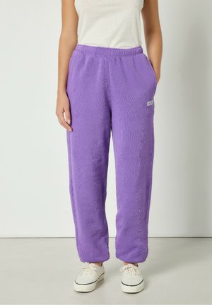 Violet vilnoniai sportiniai džinsai su elastingu juosmeniu, šoninėmis kišenėmis ir stangria tekstūra. Kairėje pusėje yra logotipas.