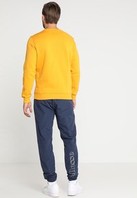 Felpa gialla con polsini e orlo a coste, abbinata a pantaloni da jogging blu navy con stampa del logo bianca. Sneakers bianche completano il look.
