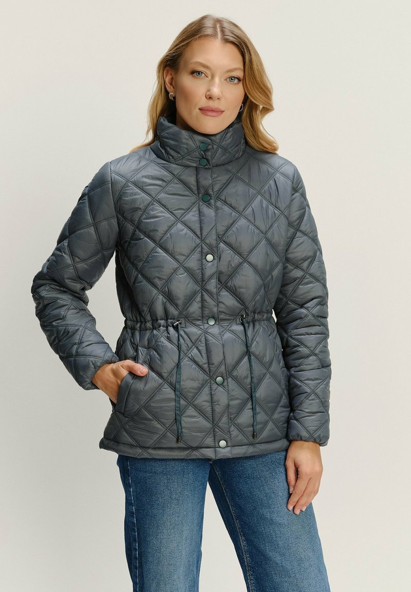 Žena v sivej prešívanej bunde so zapínaním na cvočky a šnúrkami v páse, v kombinácii s modrými džínsami, stojí pred jednoliatym pozadím.