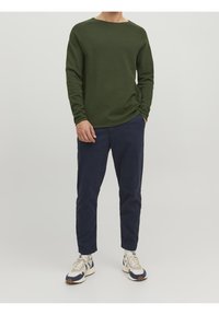 Olivgrön långärmad stickad tröja med vid halsringning, kombinerad med mörkblå byxor och sportskor med en design i vitt och mörkblått.