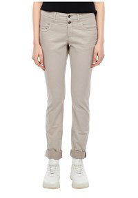 Personne portant un pantalon beige retroussé, des baskets blanches et un haut noir, debout avec les mains détendues le long du corps.
