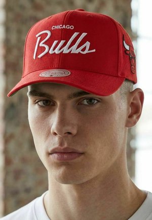 Young man wearing a red Chicago Bulls cap with logo on the side, neutral expression, beige brick wall background.