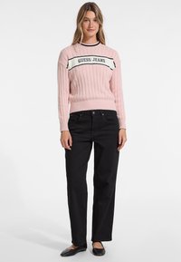 Rosa ribbad tröja med svart och vit horisontell rand som har "GUESS JEANS"-logotypen, ihop med svarta raka byxor och platta skor.