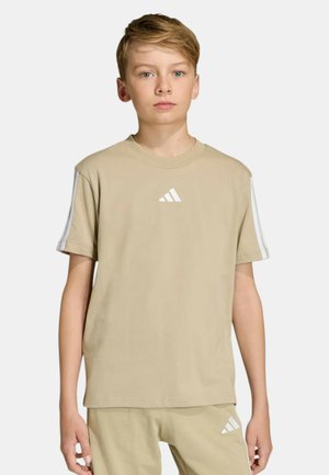 Ragazzo biondo che indossa una t-shirt beige Adidas e pantaloncini con strisce bianche e logo, in piedi davanti a uno sfondo chiaro e uniforme.