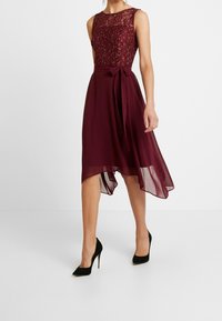 Ärmlös burgundy-klänning med spetsöverdel, midjeband och asymmetrisk chiffongkjol, bärs med svarta högklackade pumpar.