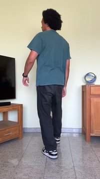 Pessoa vestindo uma t-shirt azul-água e jeans pretos, em pé sobre um chão de azulejos, de costas para um armário de madeira e uma televisão.