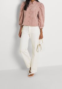 Camisa de corduroy rosa empolvado con mangas abullonadas y botones, combinada con jeans rectos blancos y un bolso mini blanco. Sandalias con tiras finas.