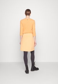 Haut orange à manches longues avec un col rond associé à une jupe orange clair. Collants gris foncé et bottines noires à semelles épaisses.