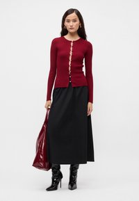 Bordo raštuotas kardiganas su mygtukų detalėmis, derinamas su juoda maksi sijonu. Aksesuarius sudaro blizgi raudona rankinė ir juodi smailiakulniai batai.