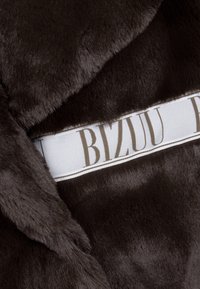 Nepvacht in donkerbruin met een zachte textuur. Heeft een witte lint met "BIZUU" in bruine letters en een omzoomde rand.