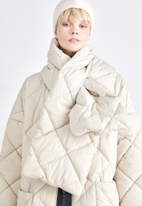 Casaco acolchoado em creme claro com colarinho alto, apresentando um design oversized e drapeado, com padrões de costura visíveis.