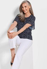 Marineblauwe blouse met stippen en korte fladdermouwen, gecombineerd met witte broeken. Model zittend, toont een relaxte pasvorm en lichtgewicht stof.
