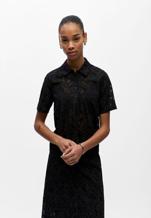 Kobieta ubrana w czarną koronkową koszulę z krótkim rękawem zapinaną na guziki oraz pasującą spódnicę, stoi złożonymi rękami na jednolitym białym tle.