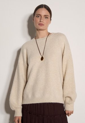 Donna con i capelli castani raccolti indossa un maglione beige peloso, gonna scura plissettata e una lunga collana con ciondolo ovale su sfondo semplice.