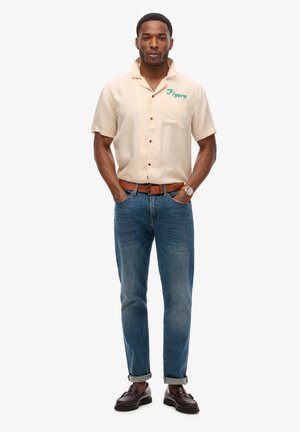 Camicia beige a maniche corte con testo verde "Flyers", due bottoni rossi e una tasca. Abbinata a jeans blu aderenti e scarpe marroni.