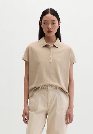 Młoda kobieta o długich ciemnych włosach, ubrana w beżową koszulkę polo z krótkim rękawem, włożoną do beżowych spodni z wysokim stanem, stojąca na jednolitym białym tle.