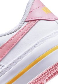 Zapatilla blanca con acentos en rosa, parte superior sintética suave, franja amarilla en la entresuela y suela de goma texturizada con un patrón de dibujo.