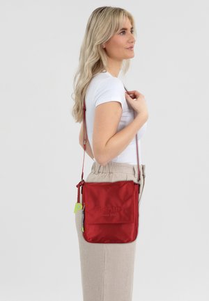 Red nylon crossbody bag with embossed brand logo, adjustable strap, and green accent tag. Smooth texture and compact rectangular shape.