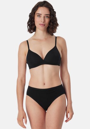 Femme aux cheveux bouclés mi-longs portant un soutien-gorge noir et une culotte taille haute noire assortie, sur un fond uni.
