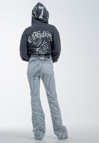 Sudadera gris con un gráfico de dragón y rosa en la parte trasera, combinada con unos jeans acampanados de color azul claro que presentan un sutil patrón de rayas de cebra y un logo bordado.