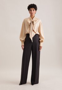 Blusa di raso beige con un grande fiocco al collo, abbinata a pantaloni scuri con vita alta e taglio elegante. Tessuto morbido, vestibilità rilassata.
