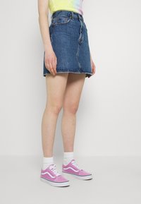 En person som bär en blå denim-minikjol, en t-shirt med tie-dye-mönster, vita strumpor och rosa Vans-sneakers står framför en enkel vit bakgrund.