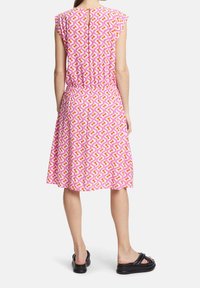 Robe rose avec un motif géométrique en jaune et orange, manches courtes, taille élastique, longueur genou, détail d'ouverture au dos, portée avec des sandales noires.