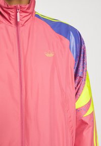 Pink windbreaker jacket with zip fastening, featuring purple and yellow accents and a subtle logo. Smooth, lightweight material with a shiny texture.